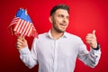 Young man with blue eyes holding flag of united states of america over red isolated background pointing and showing with thumb up Royalty Free Stock Photo