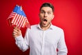 Young man with blue eyes holding flag of united states of america over red isolated background scared in shock with a surprise Royalty Free Stock Photo