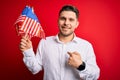 Young man with blue eyes holding flag of united states of america over red isolated background with surprise face pointing finger Royalty Free Stock Photo