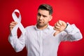 Young man with blue eyes holding white ribbon as lung cancer awareness and stop women violence with angry face, negative sign Royalty Free Stock Photo