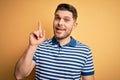 Young man with blue eyes wearing casual striped t-shirt over yellow background pointing finger up with successful idea Royalty Free Stock Photo