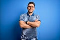 Young man with blue eyes wearing glasses and casual striped t-shirt over blue background happy face smiling with crossed arms Royalty Free Stock Photo