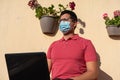 Young man with blue face mask. He is working with his laptop from the balcony of his house.