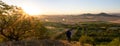 Young man in blue jacket with backpack standing on hill and looking to Czech central mountain valley at sunrise. Panoramic