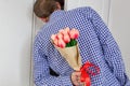 A young man in a blue plaid shirt and jeans, holding a bouquet of tulips behind his back, and peeks in the open door Royalty Free Stock Photo