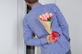 A young man in a blue plaid shirt and jeans, holding a bouquet of tulips behind his back, and peeks in the open door Royalty Free Stock Photo