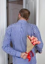 A young man in a blue plaid shirt and jeans, holding a bouquet of tulips behind his back, and peeks in the open door Royalty Free Stock Photo