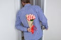 A young man in a blue plaid shirt and jeans, holding a bouquet of tulips behind his back, and peeks in the open door Royalty Free Stock Photo