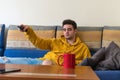 Young man, bored, watching the television while changing channels on it, laying in the sofa