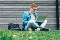 Young man businessman in shirt, jeans and glasses sitting on green grass and using laptop computer on the background of a gray Royalty Free Stock Photo