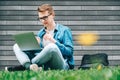 Young man businessman in shirt, jeans and glasses sitting on green grass and using laptop computer on the background of a gray Royalty Free Stock Photo