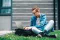 Young man businessman in shirt, jeans and glasses sitting on green grass and using laptop computer on the background of a gray Royalty Free Stock Photo