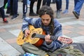 Young man busker rastaman wearing dreadlocks, sitting on pavement and playing acoustic guitar, pedestrians legs walking