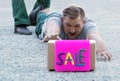 A young man buyer is lying on the sidewalk at the store and screaming with tension crawling to the box with the inscription sale.