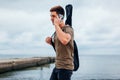 Young man carrying acoustic guitar and talking on phone walking on cloudy beach