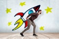 Young man in casual clothes carrying heavy colorful cartoon space rocket on his back on white wall with yellow stars Royalty Free Stock Photo