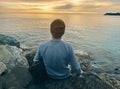 Young man in casual clothes sits on the rocks near the sea. Caucasian guy in a blue sweater and hat holds his jacket and looking Royalty Free Stock Photo