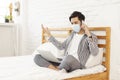 Young man in casual clothes wearing mask, sitting on bed at home office, chatting on cell phone Royalty Free Stock Photo