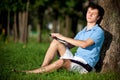 Young man sitting on green grass near tree and reading book Royalty Free Stock Photo