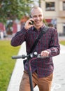 Young man in casual trousers and shirt standing next to his electric scooter, talking on the mobile phone, smiling Royalty Free Stock Photo