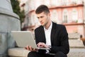 Young man in classic black suit and white shirt with wireless earphones thoughtfully working on laptop while sitting on Royalty Free Stock Photo