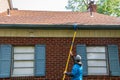 Young man cleaning the soffit of a one story house with a brush on a long pole. The house has blue shutters