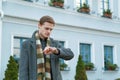 Young man in coat is looking at his watch while standing outdoors in the city. Time appointment Concept.