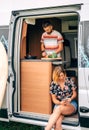 Young man cooking in a camper van while his wife looks at the mobile