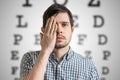 Young man is covering his face with hand and checking his vision. Chart for eye sight testing in background Royalty Free Stock Photo