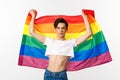 Young man in crop top, with glitter on face, raising pride flag with confident emotion. Queer person smiling with a lgbt