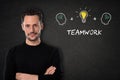 Young man with crossed arms, `Teamwork` text, heads and light bulb idea on a blackboard background. Royalty Free Stock Photo