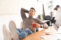 Young man with cup of drink relaxing at table in office Royalty Free Stock Photo