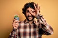 Young man with curly hair and beard holding small cactus plant pot over yellow background with happy face smiling doing ok sign Royalty Free Stock Photo