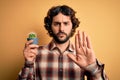 Young man with curly hair and beard holding small cactus plant pot over yellow background with open hand doing stop sign with Royalty Free Stock Photo