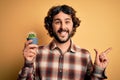 Young man with curly hair and beard holding small cactus plant pot over yellow background very happy pointing with hand and finger Royalty Free Stock Photo