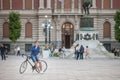 Young man, a cyclist, having a phone call sitting on a bicycle with a blurred trg republike republic square and the kod konja