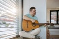 Young man with down syndrome playing acoustic guitar, sitting by window, holding and strumming guitar, making music. Royalty Free Stock Photo