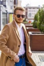 A young man dressed in a beige coat is walking down the street. A young bearded guy with a modern hairstyle and sunglasses Royalty Free Stock Photo