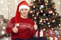 Young man dressed in retro sweater and sanat hat sit near christmas tree. New year eve, ornated fir tree. Winter holiday and love Royalty Free Stock Photo