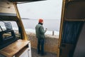 Young man drinking tea from thermos in front of camper van with a lake on background