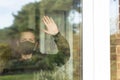 A young man with a face mask on looking and waving out the window during his self-isolation at home. Covid end of lockdown freedom