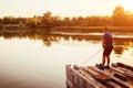 Young man fishing on river standing on bridge at sunset. Happy fiserman enjoying hobby