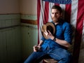 Man with beard sitting against an American flag in a dirty room