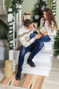 Young man and a young girl are sitting on the steps of a white staircase in a house in the eve of New Year holidays. The guy looki