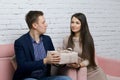 A young man gives a gift to a girl. He hands her a box of wrapping paper tied with a ribbon. Sitting on a sofa against a white Royalty Free Stock Photo