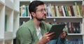 Young man in glasess sitting on the floor near shelves with book in the library, uses digital tablet computer and writes Royalty Free Stock Photo