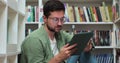 Young man in glasess sitting on the floor near shelves with book in the library, uses digital tablet computer and writes Royalty Free Stock Photo