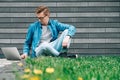 Young man with glasses businessman hipster sitting on green grass and using laptop computer on a gray wall background. Freelance Royalty Free Stock Photo