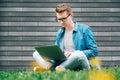Young man with glasses businessman hipster sitting on green grass and using laptop computer on a gray wall background Royalty Free Stock Photo