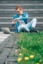 Young man with glasses businessman hipster sitting on green grass and using laptop computer on a gray wall background. Freelance Royalty Free Stock Photo